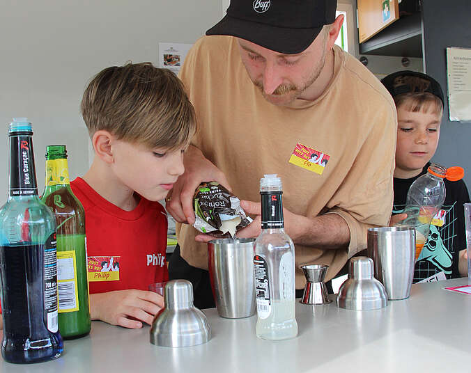 Zwei Kinder und ein Erwachsener mixen Getränke. Im Vordergrund stehen verschiedene Flaschen.